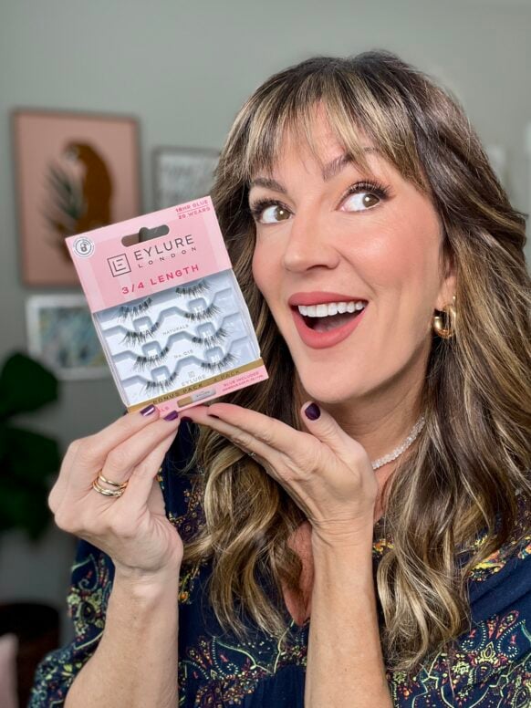 Woman with highlighted brown hair wearing navy floral top holding Eylure false lashes package, smiling at camera with natural makeup