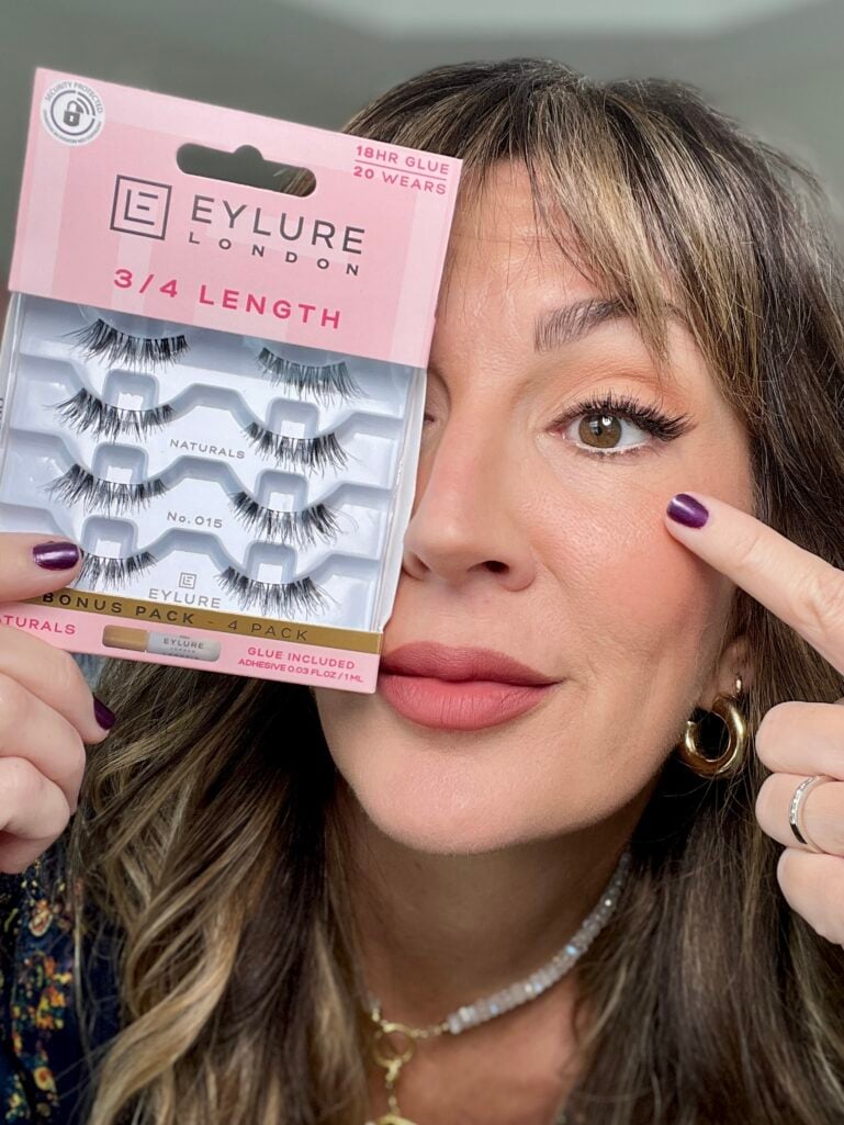 Close-up of woman wearing Eylure 3/4 length false lashes on one eye, holding lash package, showing natural-looking lash enhancement