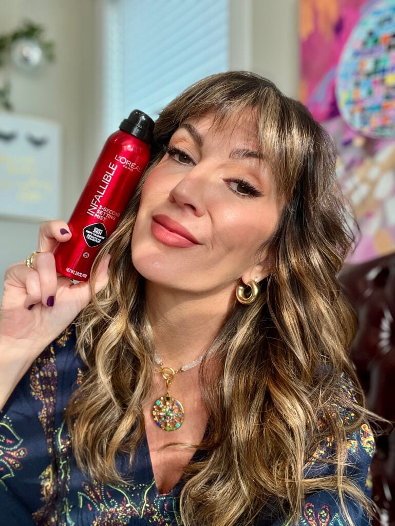Woman holding L'Oreal Infallible red makeup setting spray bottle next to face, wearing colorful pendant necklace and navy floral top
