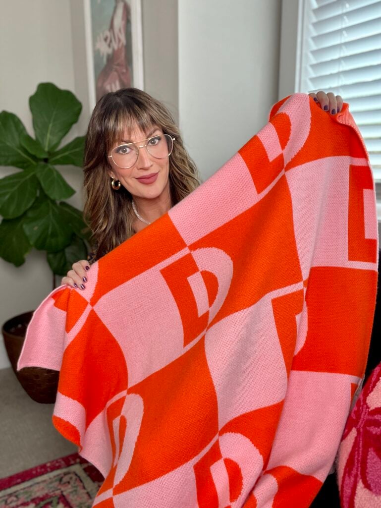 Woman wearing clear glasses wrapped in orange and pink geometric patterned Bauble Bar initial blanket in home office setting