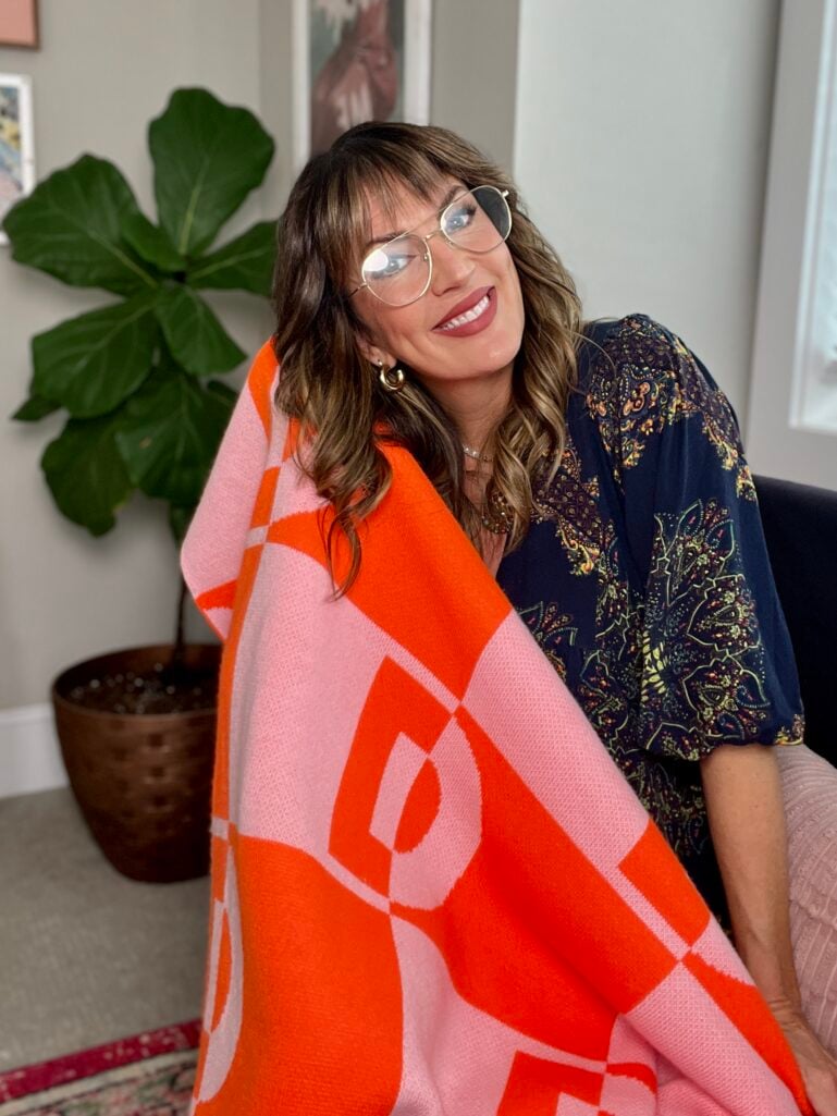 Woman smiling wrapped in orange and pink Bauble Bar monogram blanket, wearing clear aviator glasses and navy printed top