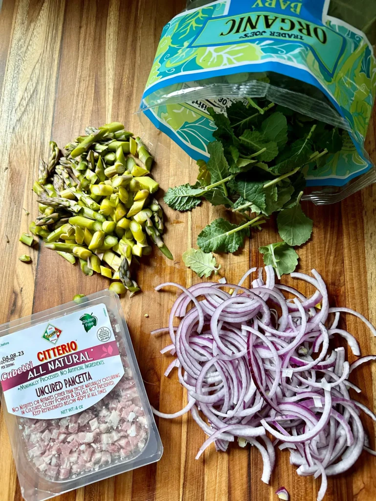 Ingredients for blue cheese bucatini on wooden cutting board: sliced asparagus, baby kale, sliced red onion, and diced pancetta