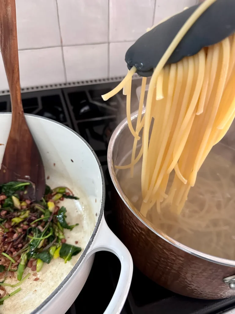 Transferring cooked bucatini pasta with tongs from pot of boiling water to pot with vegetables