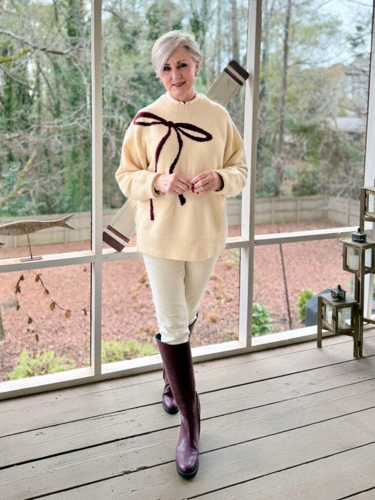 Full-length view of Beth in her cream bow sweater, ivory pants, and burgundy boots with fall foliage visible through porch windows