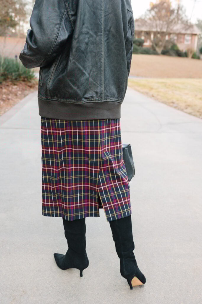 Woman over 50 styling black leather bomber jacket with tartan plaid pencil skirt and black suede heeled boots for a dressed-up holiday look