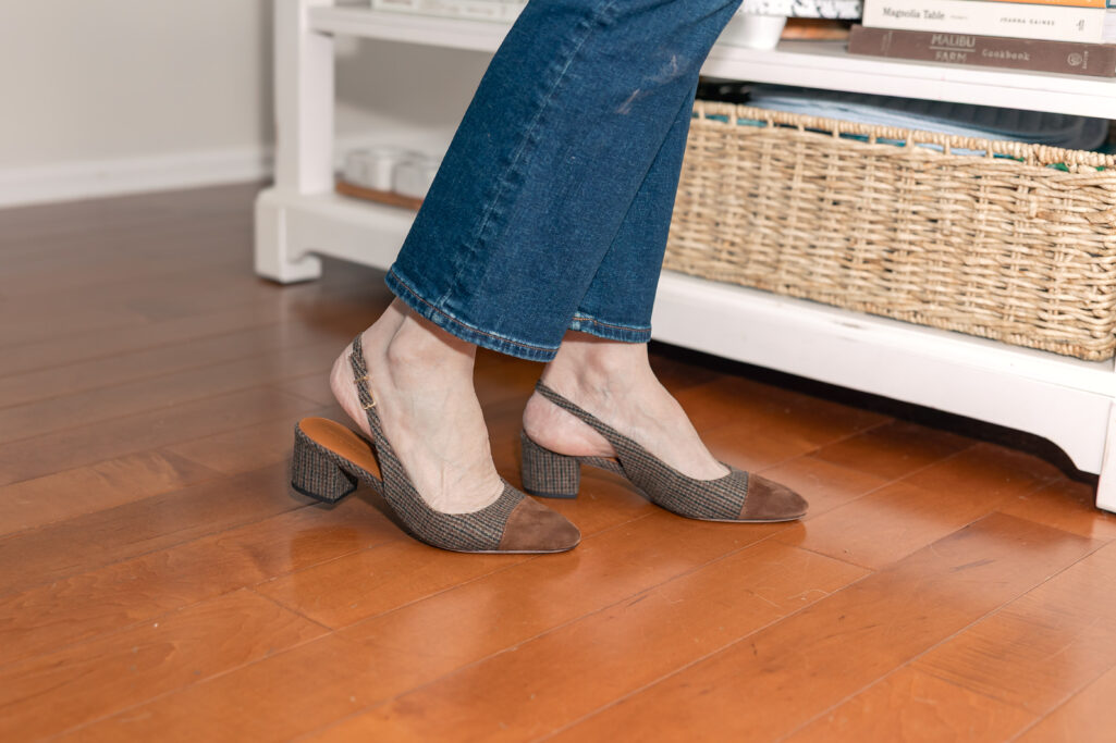 Woman over 50 wearing camel sherpa jacket over cream turtleneck sweater with dark wash jeans and tweed slingback heels holding brown suede tote in kitchen with Christmas decorations