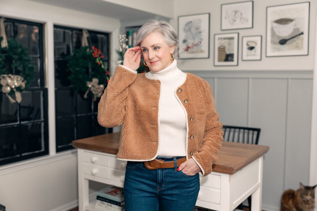 Woman over 50 wearing camel sherpa jacket over cream turtleneck sweater with dark wash jeans and tweed slingback heels holding brown suede tote in kitchen with Christmas decorations