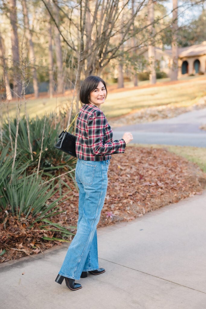 Side view of Kelly walking in Stewart tartan jacket, black sweater, and vintage flare jeans with black crossbody bag showing casual holiday outfit movement for petites