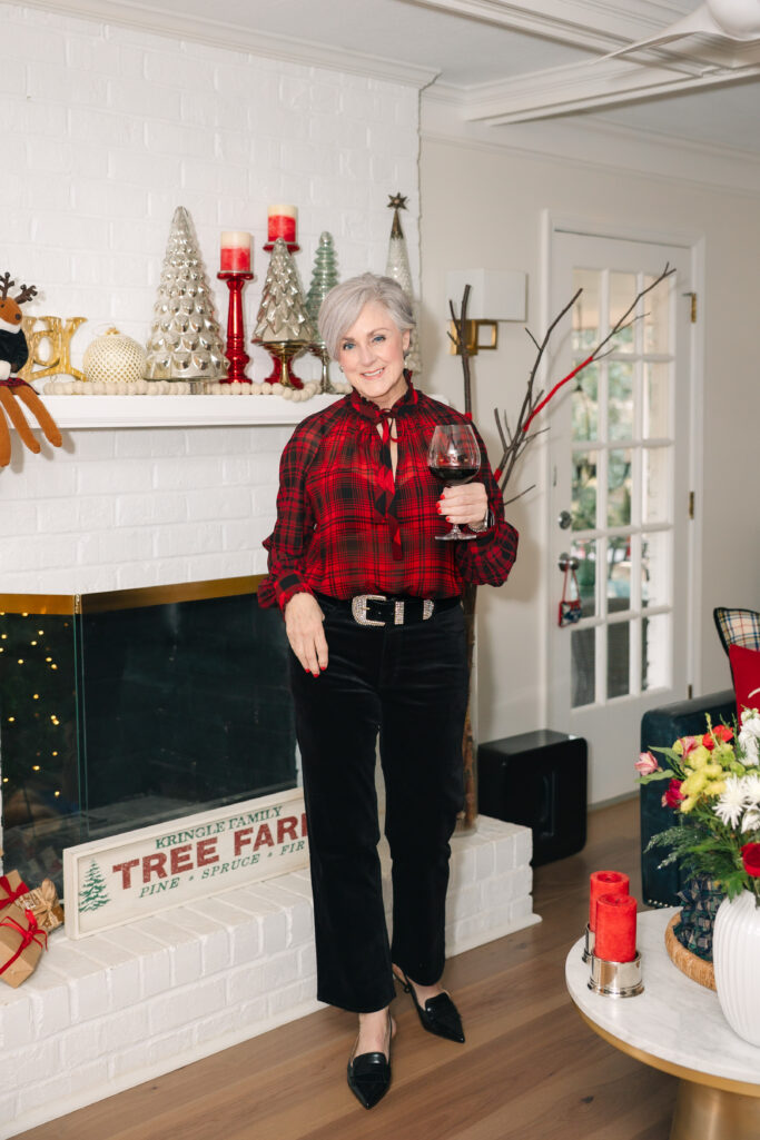 Woman over 50 wearing red and black plaid tie-neck blouse with black velvet jeans and black slingback heels standing by decorated fireplace mantel holding glass of red wine