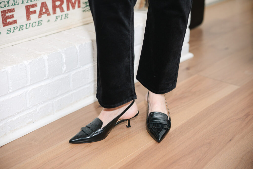 Woman over 50 wearing red and black plaid tie-neck blouse with black velvet jeans and black slingback heels standing by decorated fireplace mantel holding glass of red wine