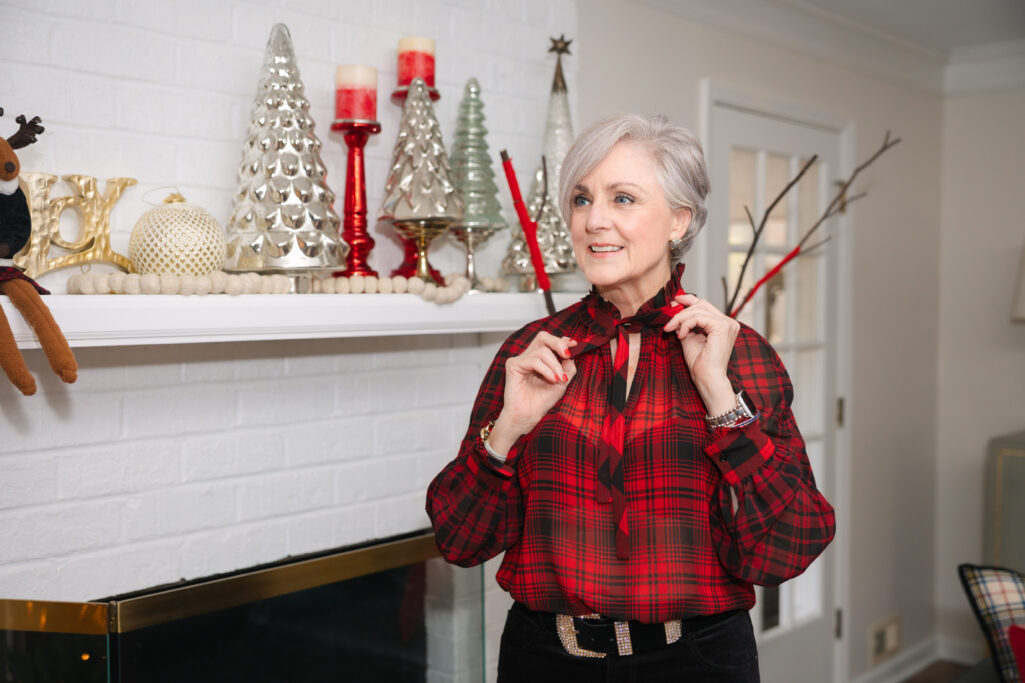 Woman over 50 wearing red and black plaid tie-neck blouse with black velvet jeans and black slingback heels standing by decorated fireplace mantel holding glass of red wine