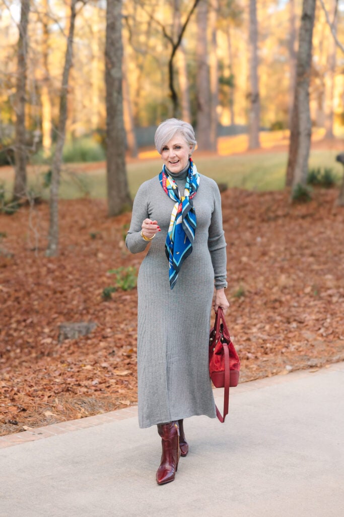 Woman over 50 wearing gray ribbed turtleneck sweater dress with colorful silk scarf, burgundy ankle boots, red leather handbag, and gold bangle bracelets