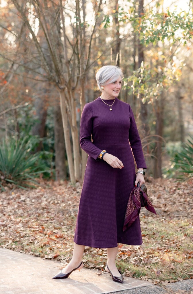 Woman over 50 wearing long-sleeve plum quilted midi dress with A-line skirt, gold heart necklace, geometric bracelet, burgundy kitten heels and croc-embossed handbag