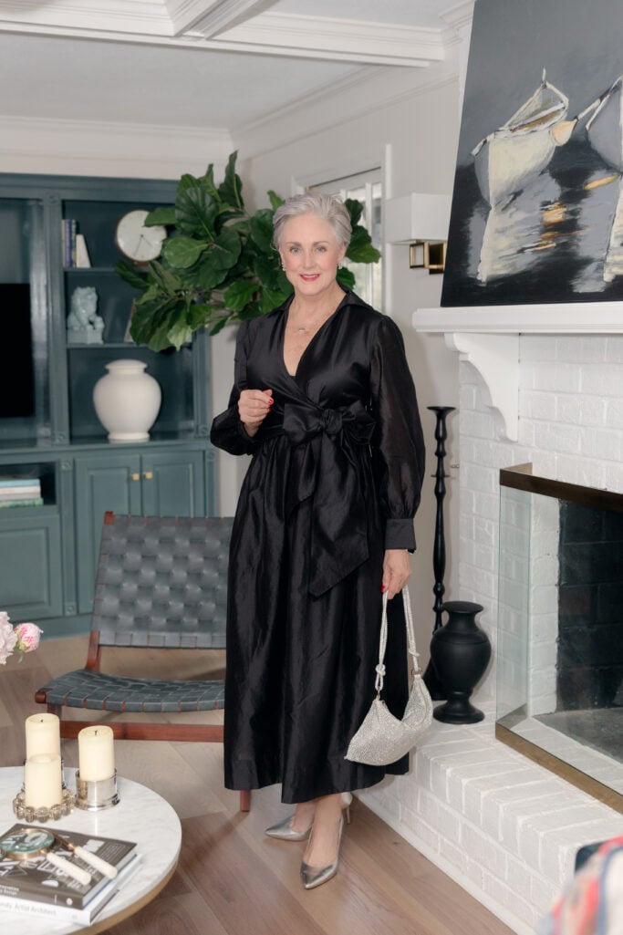 Beth Djalali wearing a black taffeta wrap dress with sheer balloon sleeves and bow waist detail, styled with silver metallic pumps and crystal mesh evening bag for a winter wedding guest look