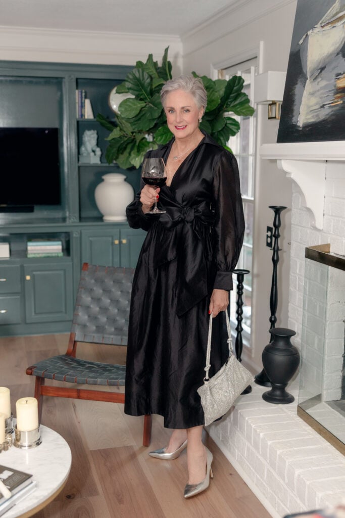 Beth Djalali wearing a black taffeta wrap dress with sheer balloon sleeves and bow waist detail, styled with silver metallic pumps and crystal mesh evening bag for a winter wedding guest look