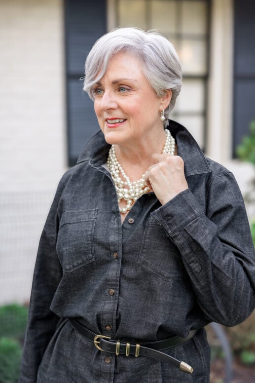 Woman over 50 with silver-gray pixie hairstyle wearing black denim shirt jacket with multi-strand pearl necklace and black leather belt, smiling outdoors with blurred house exterior in background.