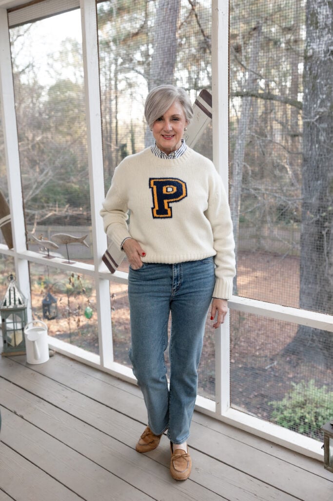 Beth smiling in a cream varsity-style sweater with navy and gold P, striped button-up collar visible, slim jeans, and tan suede loafers