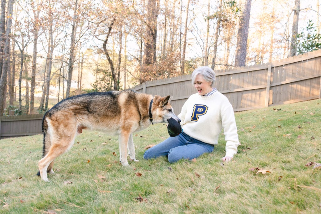 Oscar nuzzling Beth's hand as she sits on the grass in her cream letter sweater