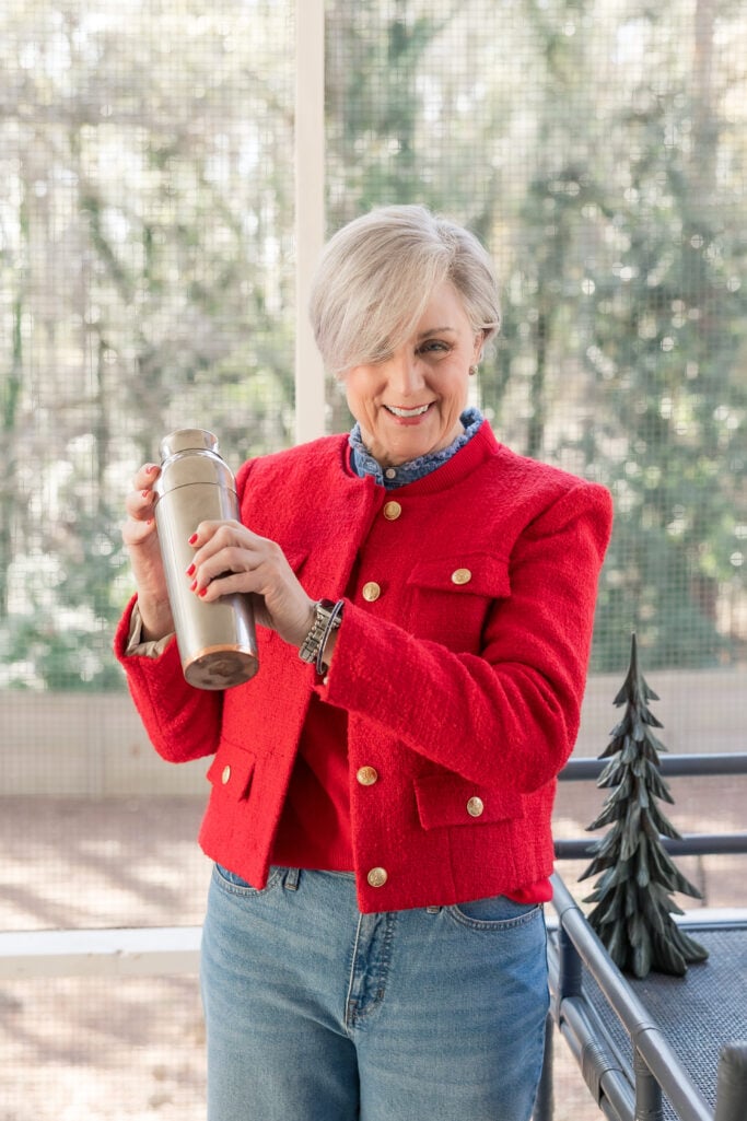 Woman with short silver-blonde hair wearing bright red textured bouclé jacket with gold buttons over denim ruffle top and light wash jeans, holding cocktail shaker on screened porch with Christmas decor