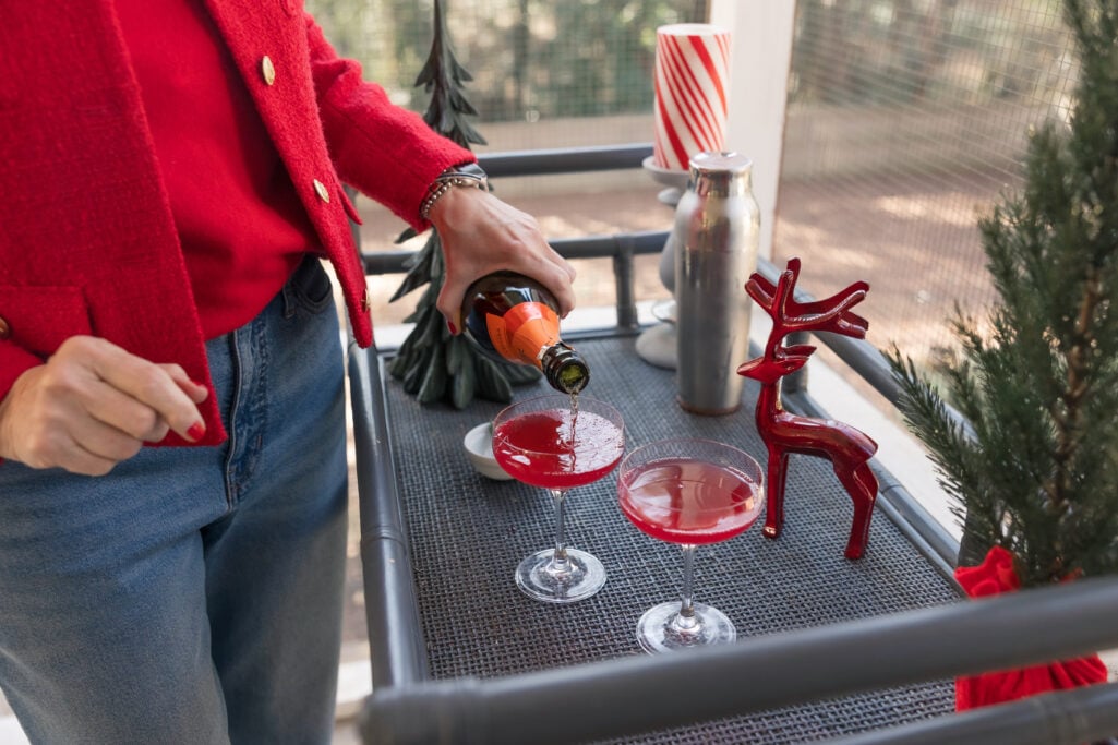 Close-up of hands in red jacket pouring ruby-red cocktail from Mionetto Prosecco bottle into coupe glass on outdoor bar with holiday decorations and candy cane striped candle
