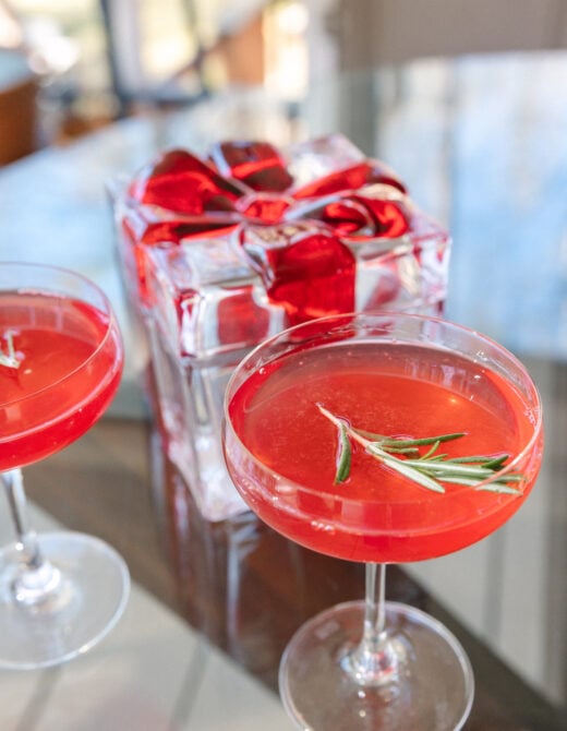 Two ruby-red Mistletoe Martinis in coupe glasses garnished with fresh rosemary sprigs, with festive wrapped gift box with red plaid ribbon in background
