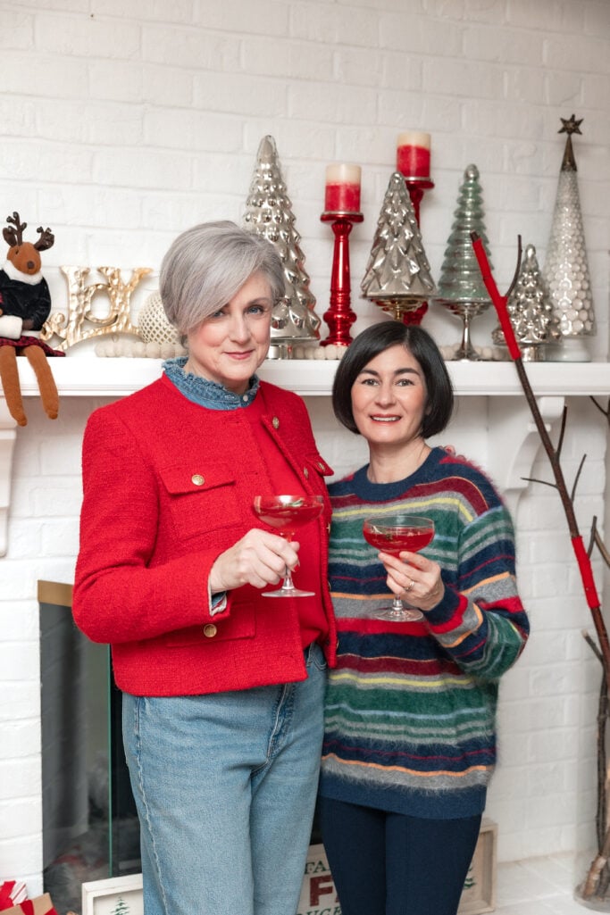 Two women standing together in front of white brick fireplace with silver Christmas trees and holiday decor, raising ruby-red cocktails in a toast, one in red bouclé jacket, one in multicolor striped sweater
