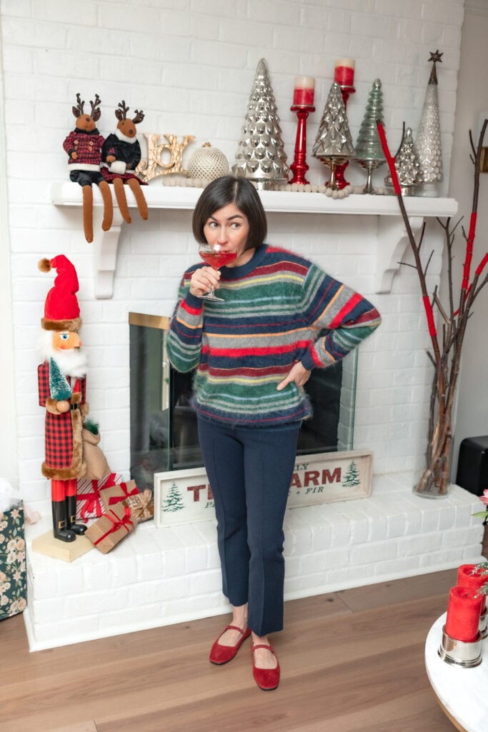 Woman with dark bob haircut in multicolor striped cashmere sweater and navy pants sipping ruby-red cocktail from coupe glass, standing in front of white brick fireplace with large nutcracker and holiday decor