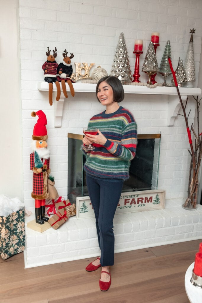 Woman in festive vintage-stripe brushed cashmere sweater in navy, green, red, yellow, and gray, navy kickout sweater pants, and red ballet flats, holding cocktail and smiling in front of decorated Christmas fireplace