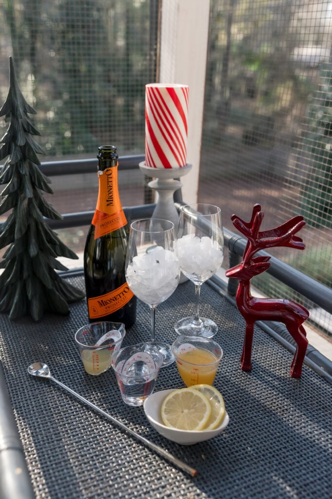 Prosecco punch ingredients including Mionetto Prosecco, wine glasses filled with ice, lemon slices, measuring cups, and bar spoon on a festive holiday table with green tree and red reindeer decor