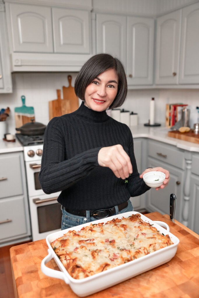Kelly sprinkling fresh herbs over the finished ham and Gruyère croissant strata in a white baking dish in the kitchen