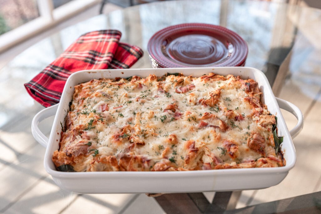 Golden-brown ham and Gruyère strata in a white baking dish with fresh herb garnish, red plaid napkin and holiday plate alongside