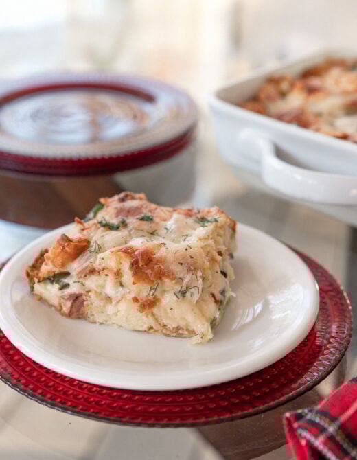 A single serving of the croissant strata on a white plate atop a red charger, showing the fluffy interior with greens and ham