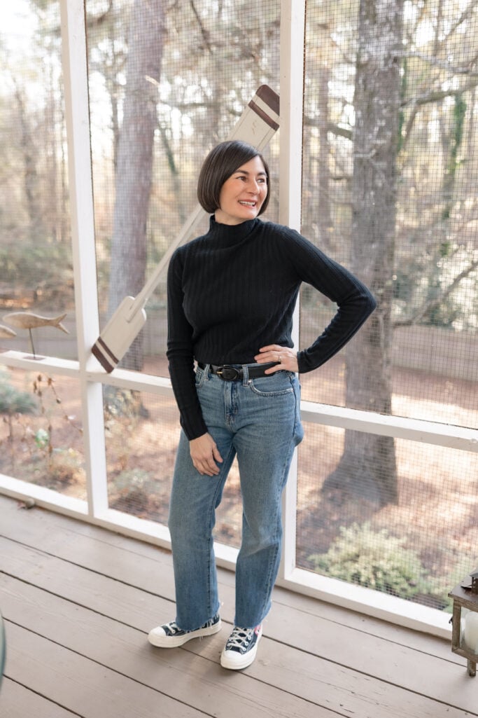 Kelly wearing a black ribbed cashmere turtleneck, straight leg jeans, and Comme des Garçons PLAY x Converse high tops on a screened porch
