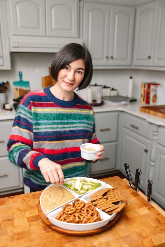 Woman with dark bob haircut wearing multicolor striped cashmere sweater in navy, green, red, and gold, holding small white bowl while arranging gingerbread cheesecake dip platter in bright white kitchen