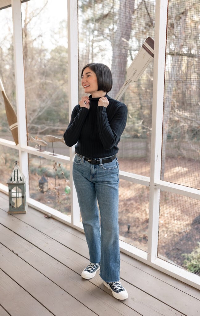 Kelly adjusting the collar of her black cashmere turtleneck, full-length view showing jeans and heart-logo Converse sneakers