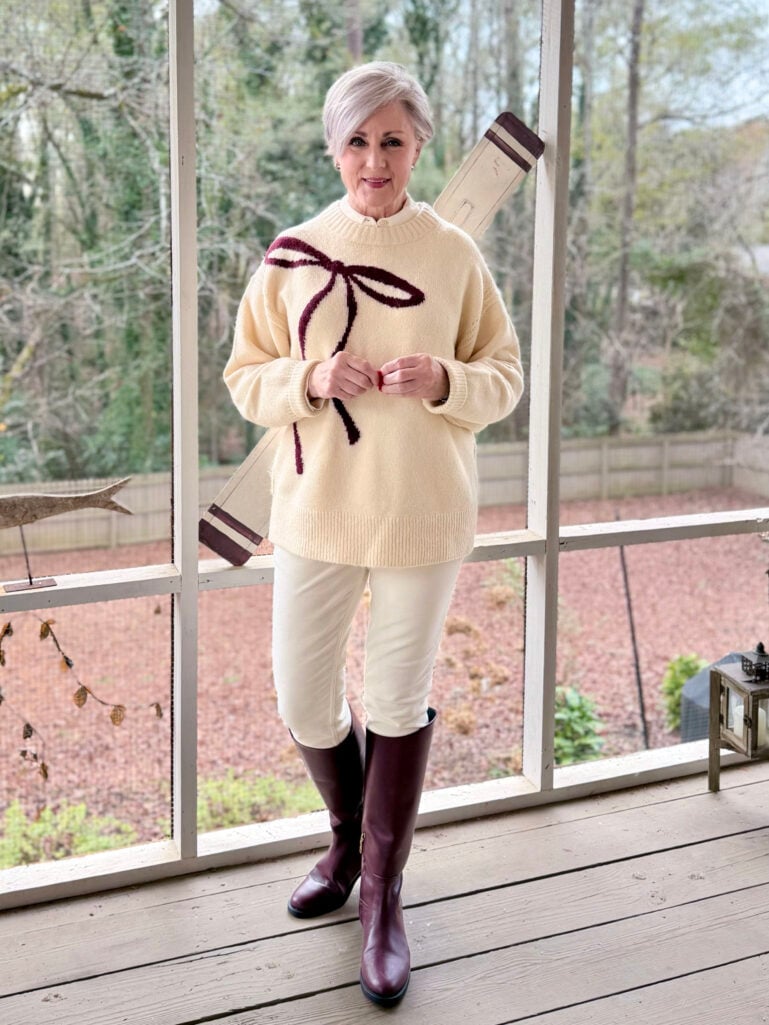Beth wearing a cream bow-print sweater with burgundy ribbon detail, ivory velveteen pants, and burgundy riding boots on a screened porch