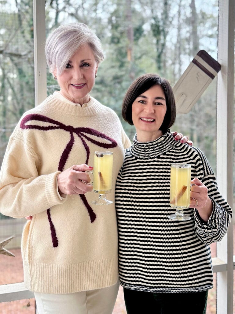 Beth and Kelly raising hot toddies in glass mugs, Beth in her bow sweater and Kelly in a black and white striped turtleneck