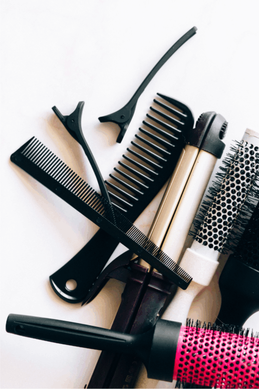Flat-lay of hair styling tools including combs, sectioning clips, a curling iron, and round brushes arranged on a white surface.