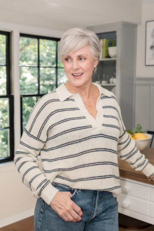 Woman with short silver hair wearing a cream and navy striped knit sweater and jeans, smiling in a bright kitchen with large windows and natural light.