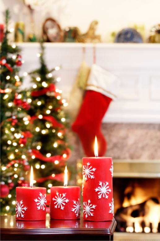 Festive holiday mantel decorated with glowing red pillar candles featuring white snowflake designs, a lit Christmas tree, hanging stockings, and a cozy fireplace creating a warm seasonal atmosphere.