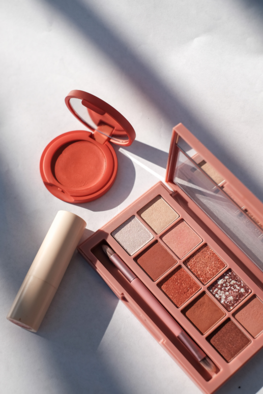 lat lay of luxury makeup products including an eyeshadow palette, cream blush compact, lipstick, and makeup brush, arranged in warm neutral tones with soft natural lighting.