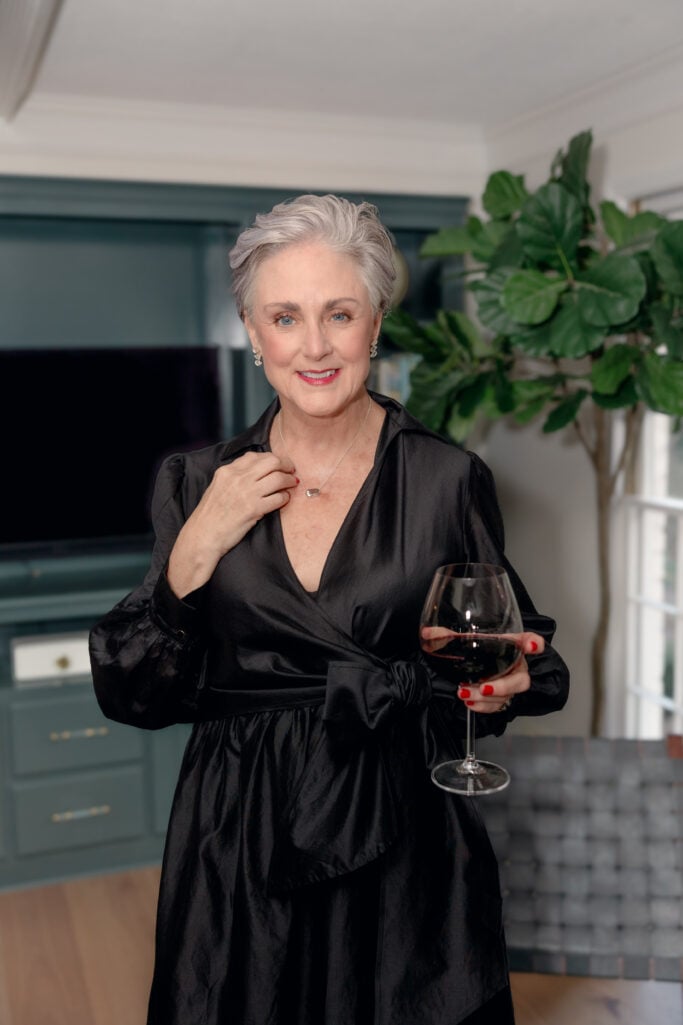 Woman over 50 wearing black taffeta wrap dress holding wine glass, showing elegant neckline and sleeve detail