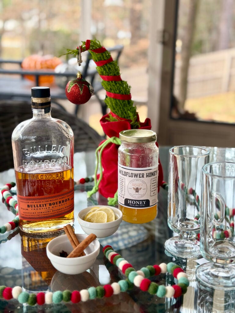 Hot toddy ingredients including Bulleit Bourbon, wildflower honey, lemon slices, cinnamon sticks, star anise, and glass mugs