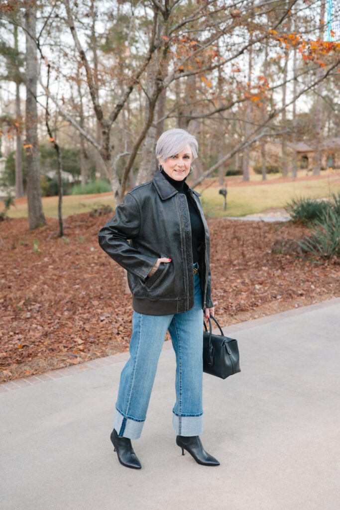 Woman over 50 wearing black oversized leather bomber jacket with black turtleneck and blue jeans, holding black leather bag