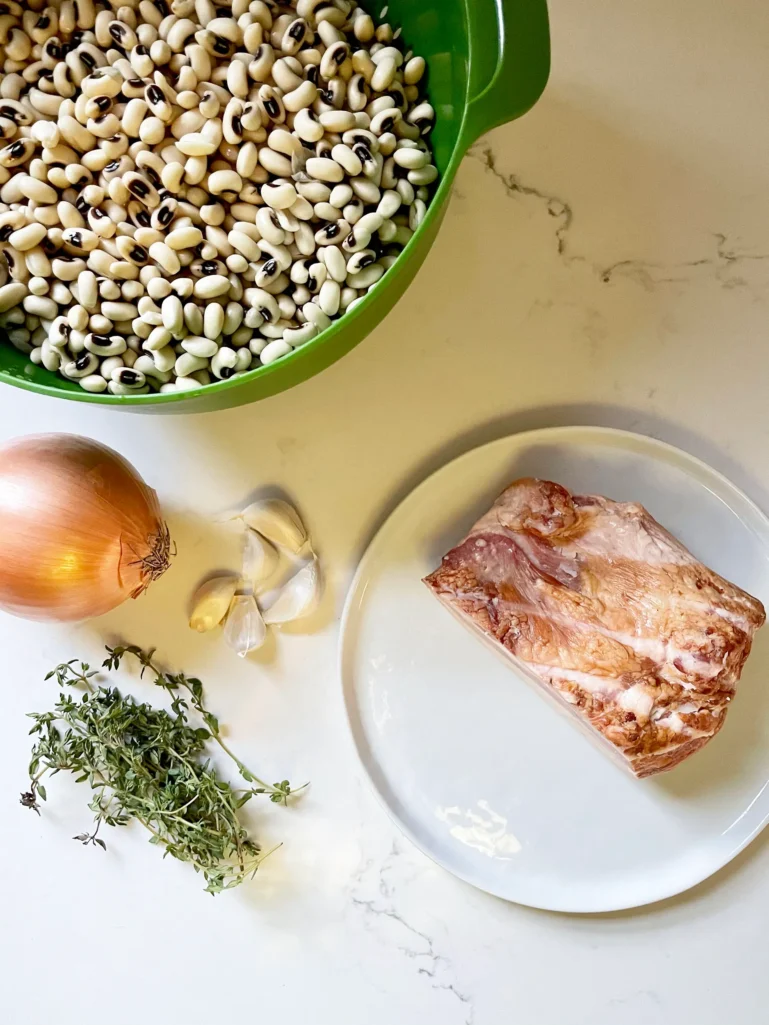 Ingredients for black-eyed peas: soaked peas in a green bowl, yellow onion, garlic cloves, fresh thyme, and slab bacon