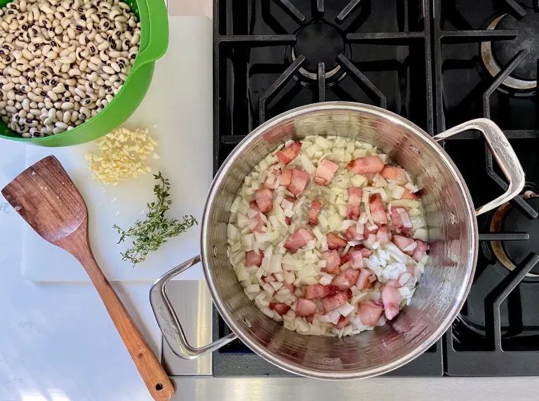 Overhead view of pot with bacon and onions on stovetop next to cutting board with soaked black-eyed peas, chopped garlic, and thyme