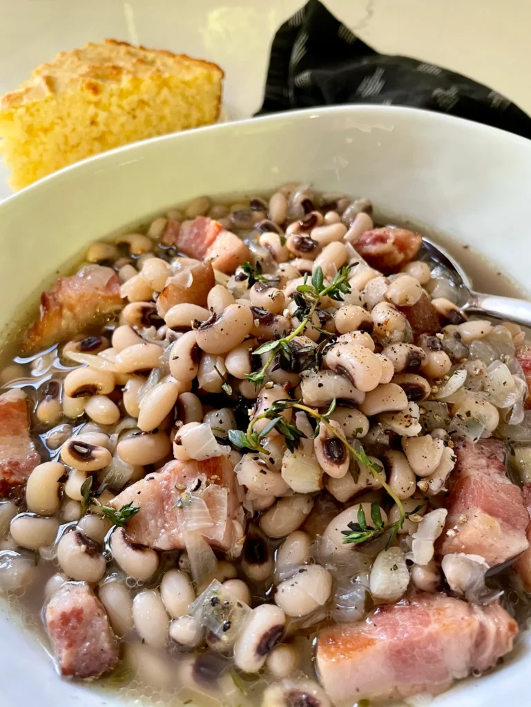 Bowl of black-eyed peas with bacon and fresh thyme served with a piece of cornbread on the side