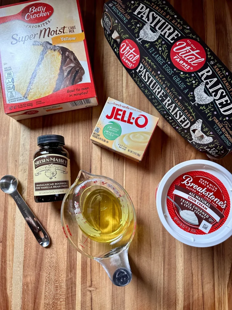 Coffee cake ingredients on wooden cutting board including yellow cake mix, pudding mix, sour cream, eggs, oil, and vanilla