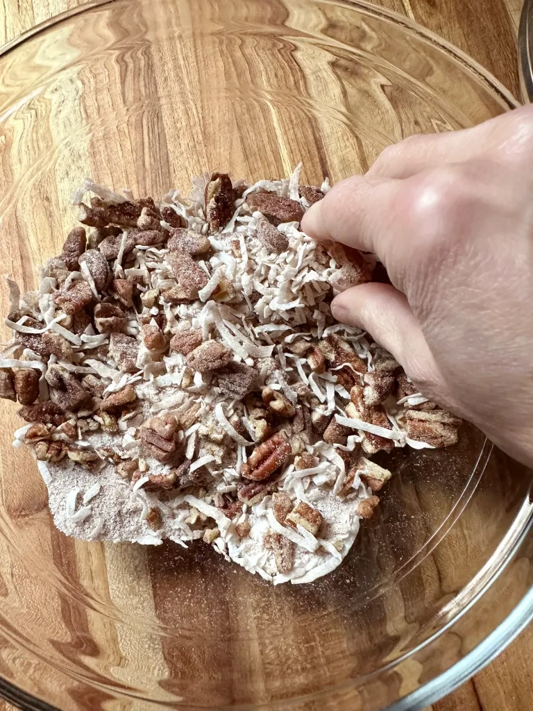 Hands mixing chocolate chips, coconut, pecans, and cinnamon sugar in a glass bowl