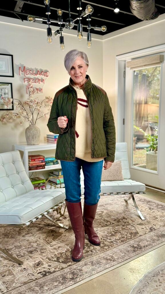 Woman over 50 wearing olive quilted field jacket with leather trim, cream cricket sweater with burgundy stripe, jeans, and burgundy riding boots in Ralph Lauren countryside style